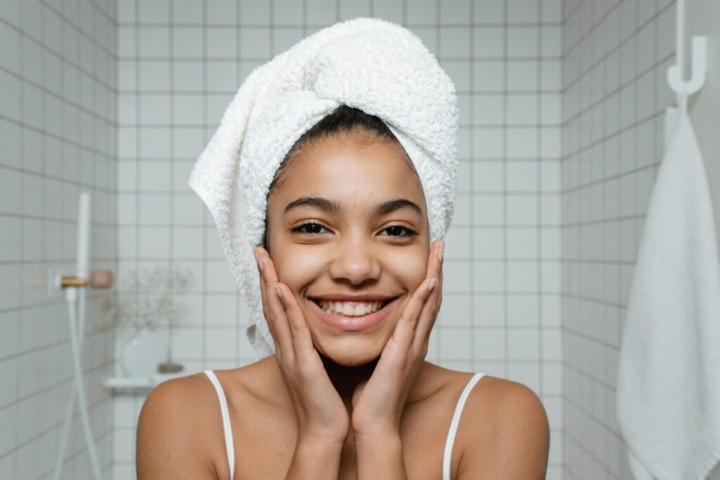 A young woman smiles during her morning skincare routine with a towel around her head.